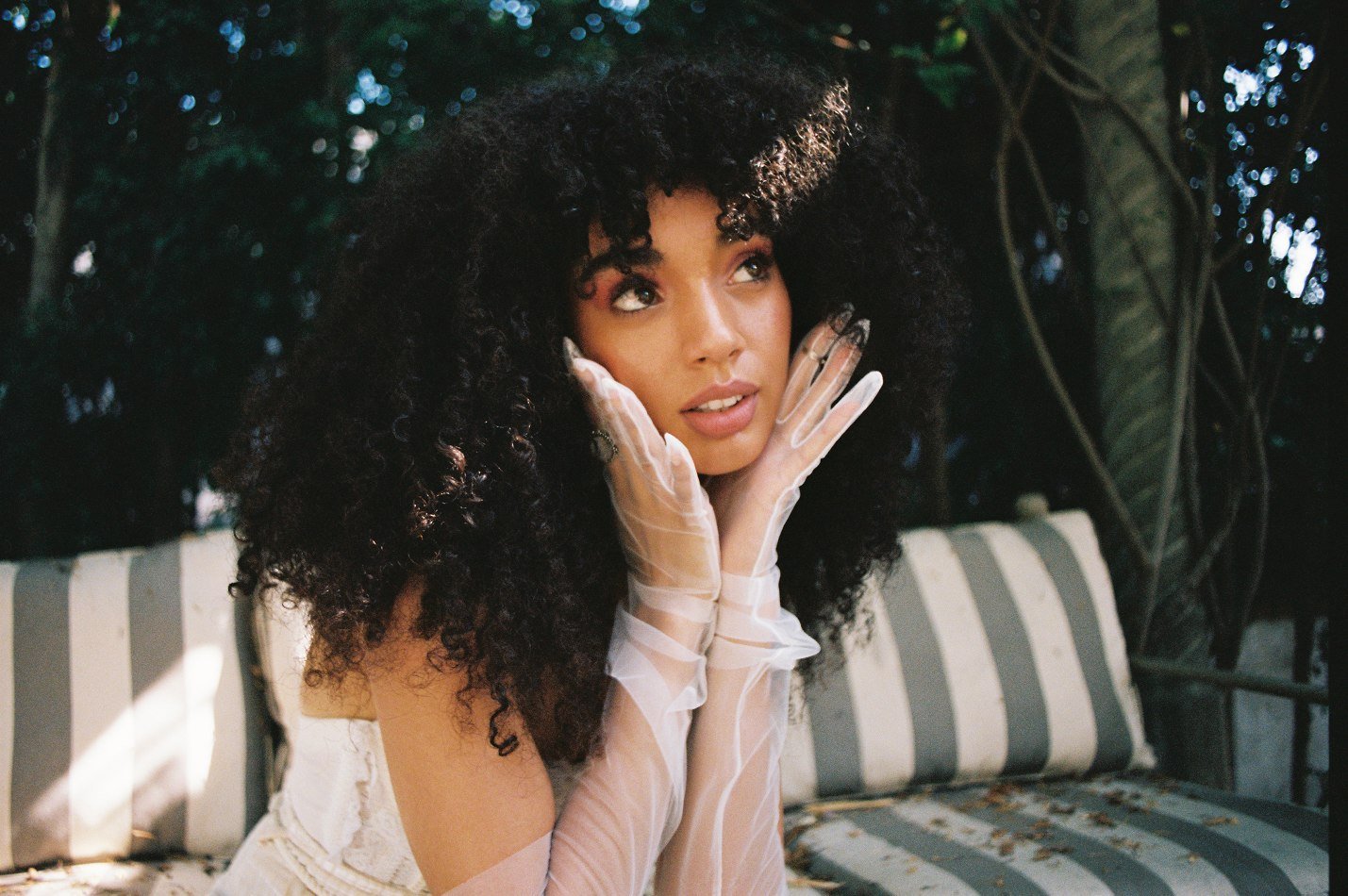 Outdoor portrait of a dark-haired woman in sheer gloves, seated on a gray striped bench with a dreamy expression, surrounded by foliage.