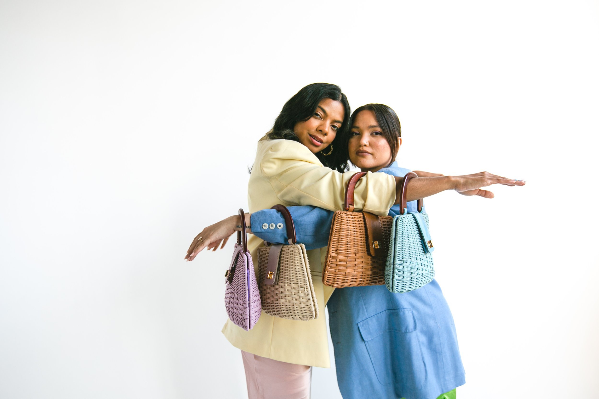 Two women pose in front of a white background, each holding multiple colorful, woven handbags with top handles. Fashion shot.