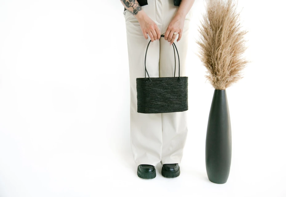 Adult is carrying a black woven handbag in a white indoor studio setting with a tall decorative tan grass arrangement nearby.