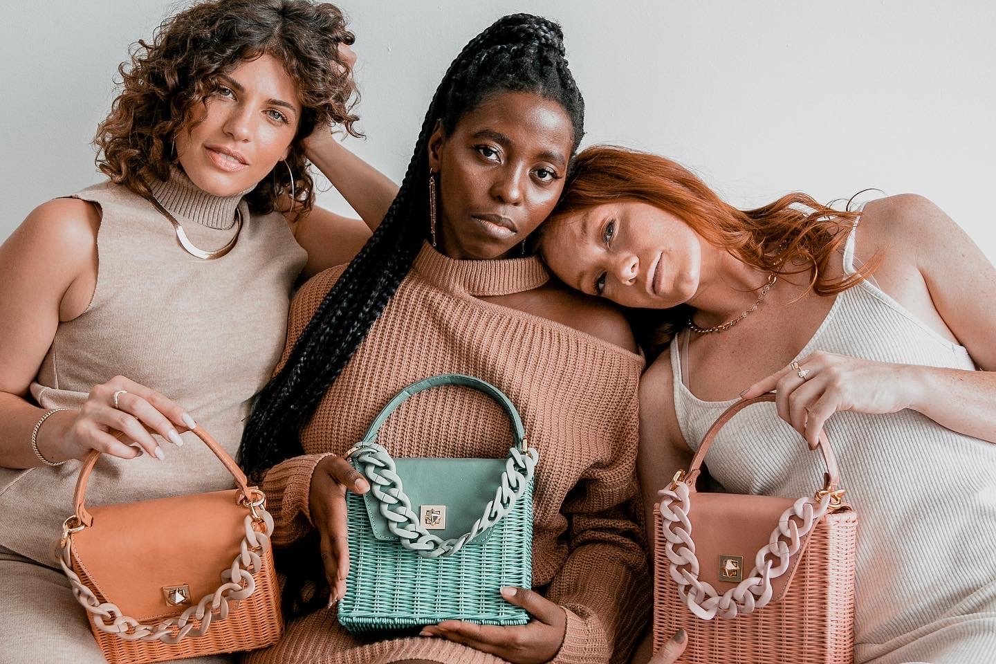 Three women pose indoors with rattan handbags, displaying neutral-tone clothing and accessories in a fashion-forward studio setting.