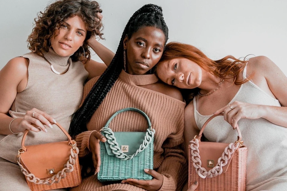 Three women pose indoors with rattan handbags, displaying neutral-tone clothing and accessories in a fashion-forward studio setting.