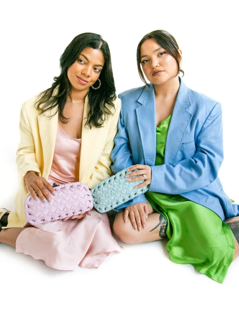 Two women in oversized blazers holding woven clutches against a white backdrop, showcasing a fashion-forward aesthetic and pastel color palette.