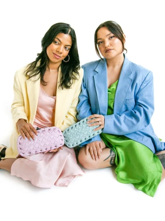 Two women in oversized blazers holding woven clutches against a white backdrop, showcasing a fashion-forward aesthetic and pastel color palette.