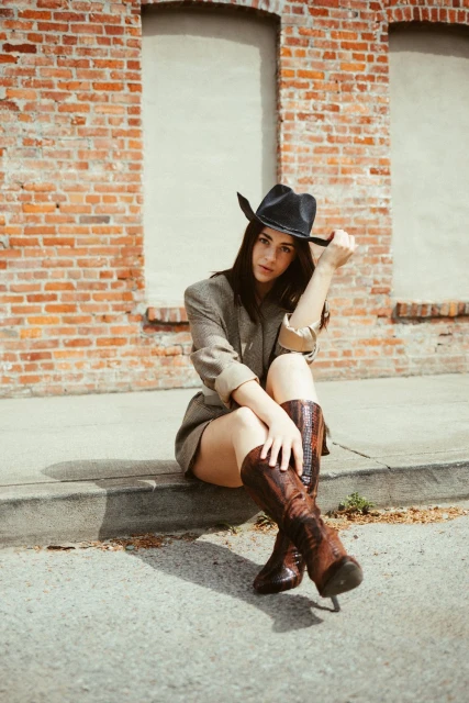 Young woman poses outdoors wearing a cowboy hat, blazer, and boots, seated near a brick wall with a concrete curb.