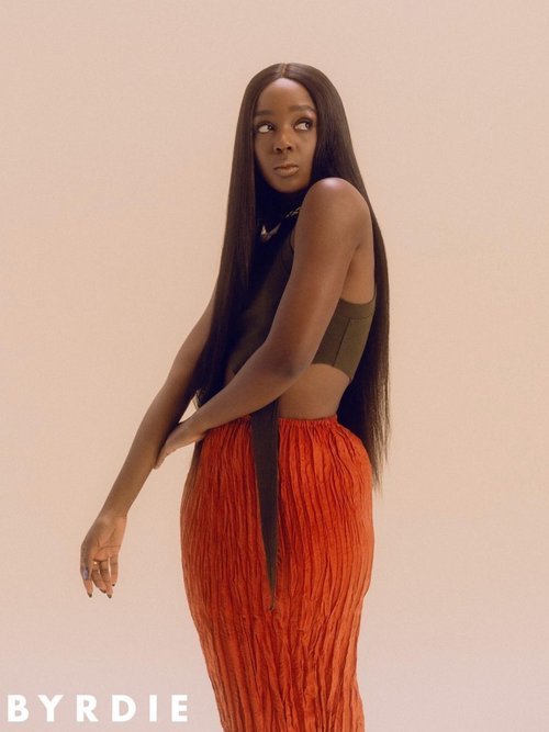 Portrait of a Black female adult model with long black hair, wearing a crop top and orange skirt, in a photo studio setting.