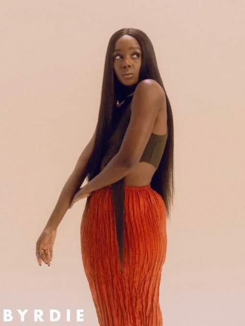 Portrait of a Black female adult model with long black hair, wearing a crop top and orange skirt, in a photo studio setting.