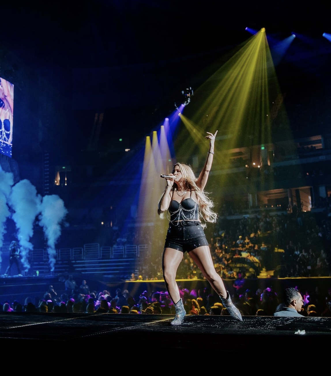 Female singer performs on stage at a music concert, holding a microphone and illuminated by yellow and blue stage lights.