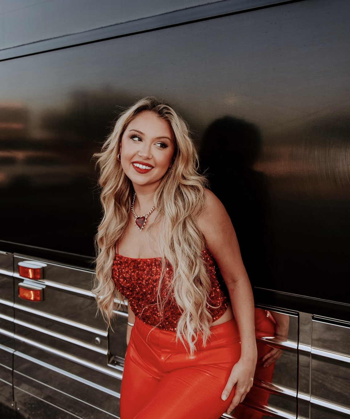 Blonde woman in red sequin top and red vinyl pants poses next to a tour bus with a sunset visible in the background.