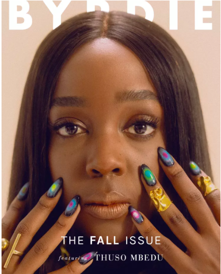 Close-up of a Black woman's face, adorned with galaxy-themed nails and gold rings, against a light beige background on a magazine cover.