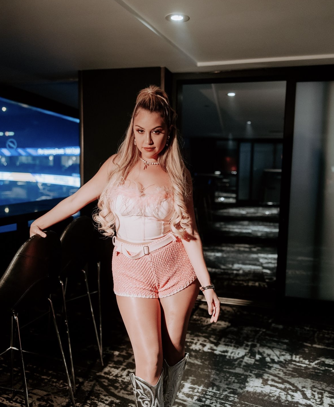 Blonde woman in pink outfit posing indoors near a glass wall and black furniture, wearing cowboy boots, with a modern aesthetic.