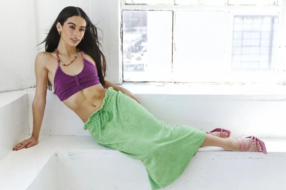 A woman with long dark hair poses indoors wearing a purple halter top and a green towel skirt, near a window with natural light.