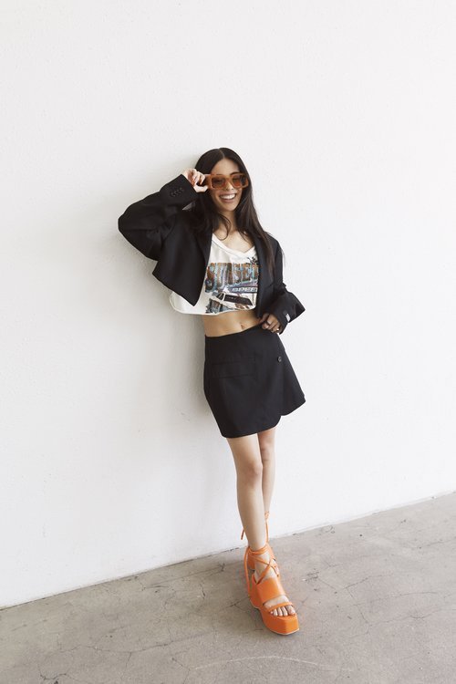 Brunette woman in black blazer and mini-skirt smiles in studio, accessorized with orange sunglasses and platform sandals.