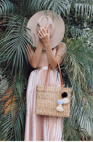 Adult woman covers face with hat, accessorized with a straw tote bag and long skirt, standing among leafy palm trees in daytime.