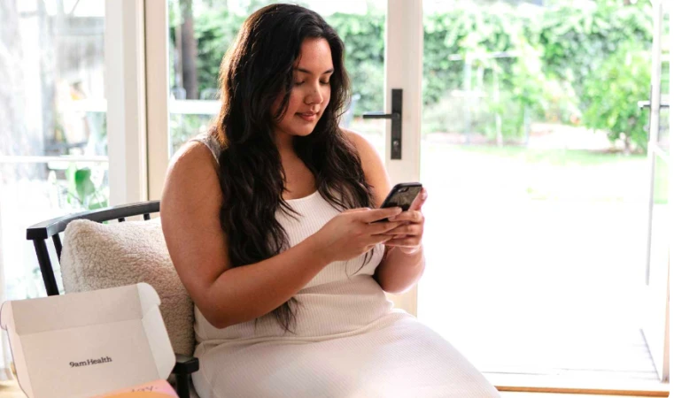 Brunette woman in a white dress sits indoors using a smartphone next to a 9am Health box, with a window and greenery visible outside.