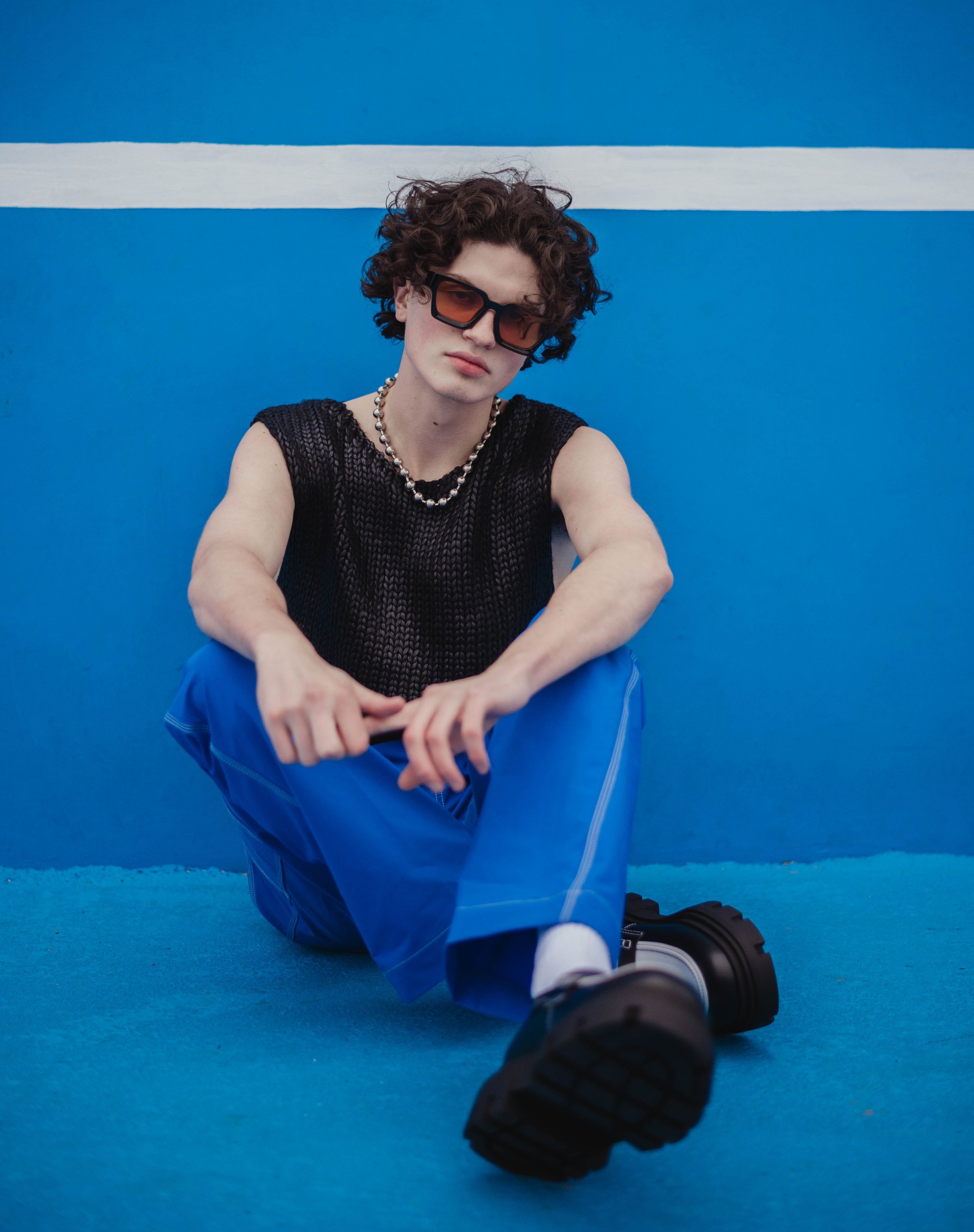 A young adult male model in an open-weave shirt and blue pants poses against a bright blue background with a white horizontal stripe.