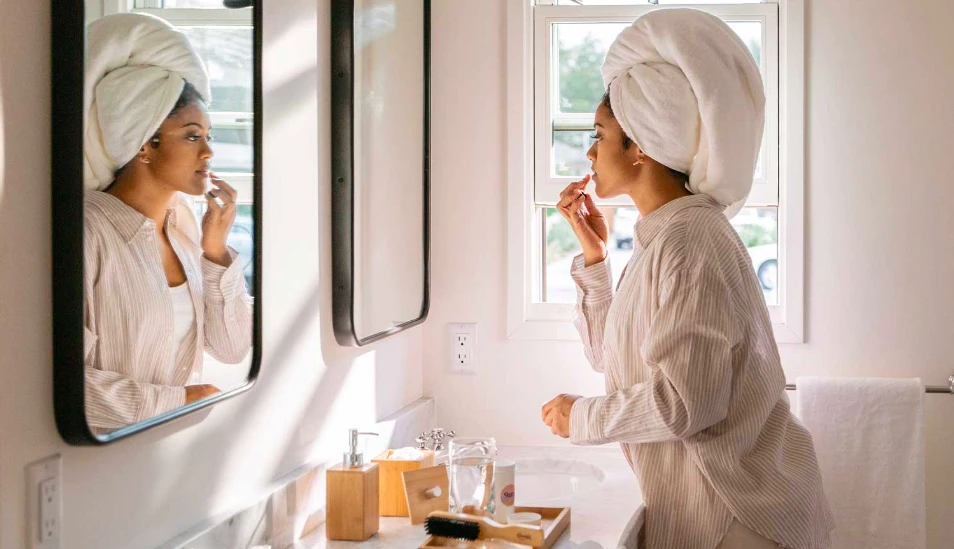 Woman in a bathroom mirror with a towel on her head, applying makeup in a brightly lit modern bathroom with white walls and marble countertop.