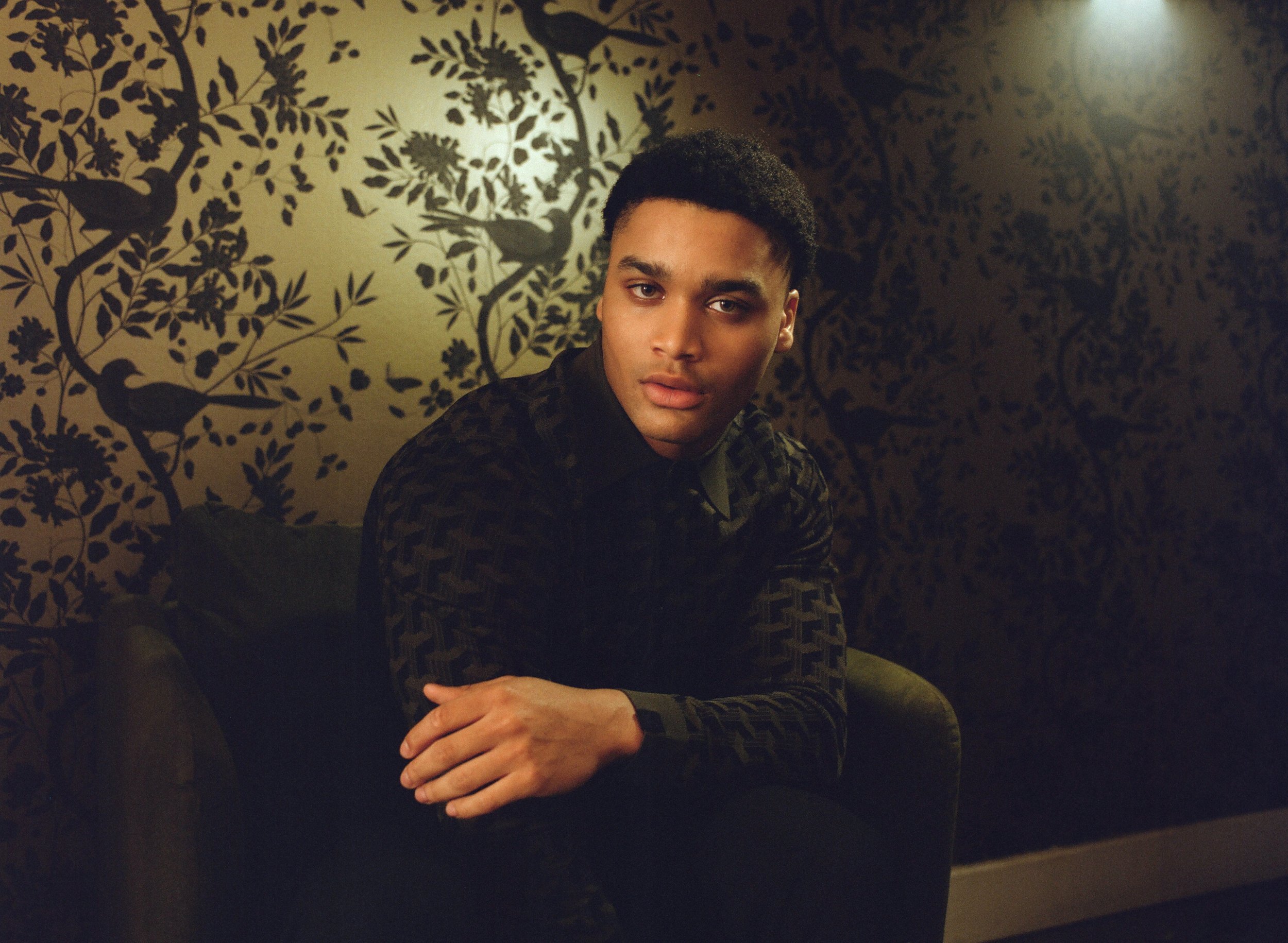 Portrait of a young adult man with dark hair and a patterned shirt, sitting in a chair in front of a floral wallpaper.