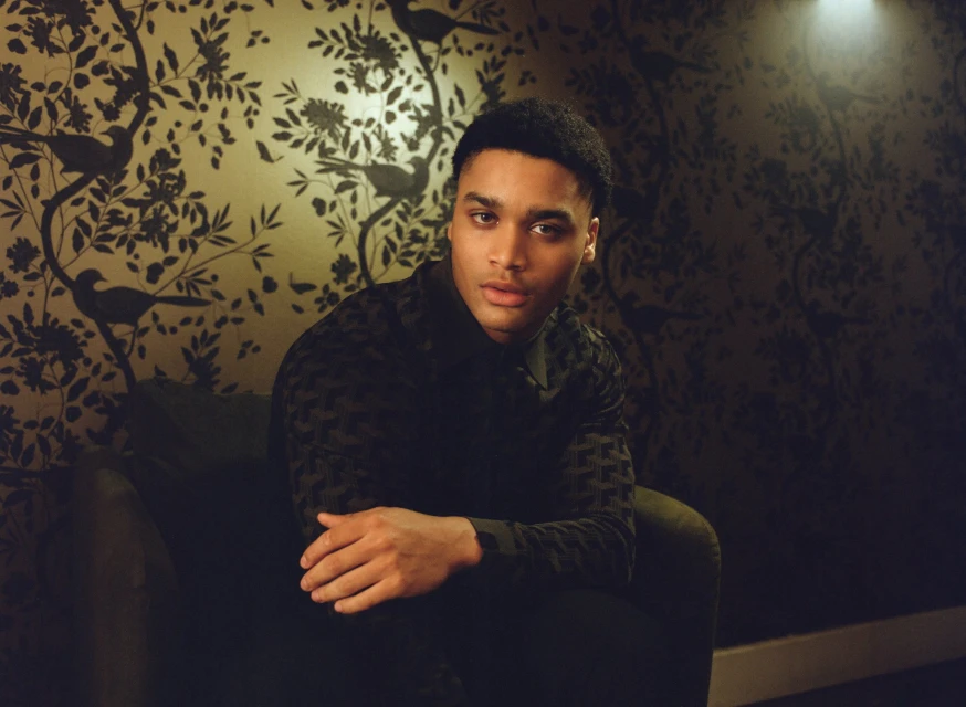 Portrait of a young adult man with dark hair and a patterned shirt, sitting in a chair in front of a floral wallpaper.