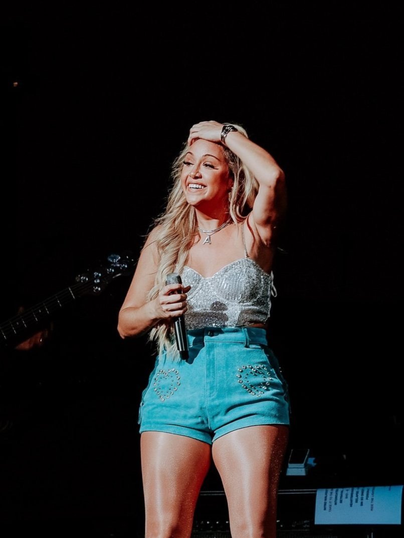 Blonde singer on stage smiles, wearing a sequined top and blue shorts, holding a microphone, performing against a dark background.