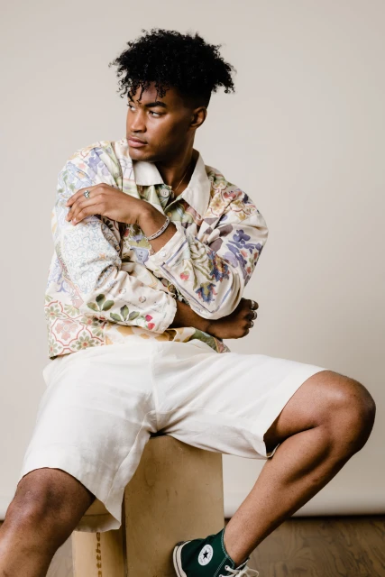 Young adult male model with curly hair sitting on a box in a patterned jacket, shorts, and sneakers in a photo studio setting.