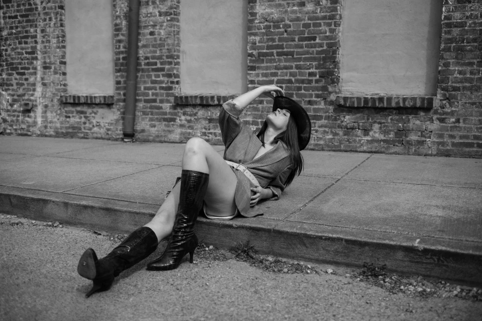 Monochrome outdoor portrait of a woman in a blazer and cowboy hat reclining on the sidewalk by a brick building in high-heeled boots.