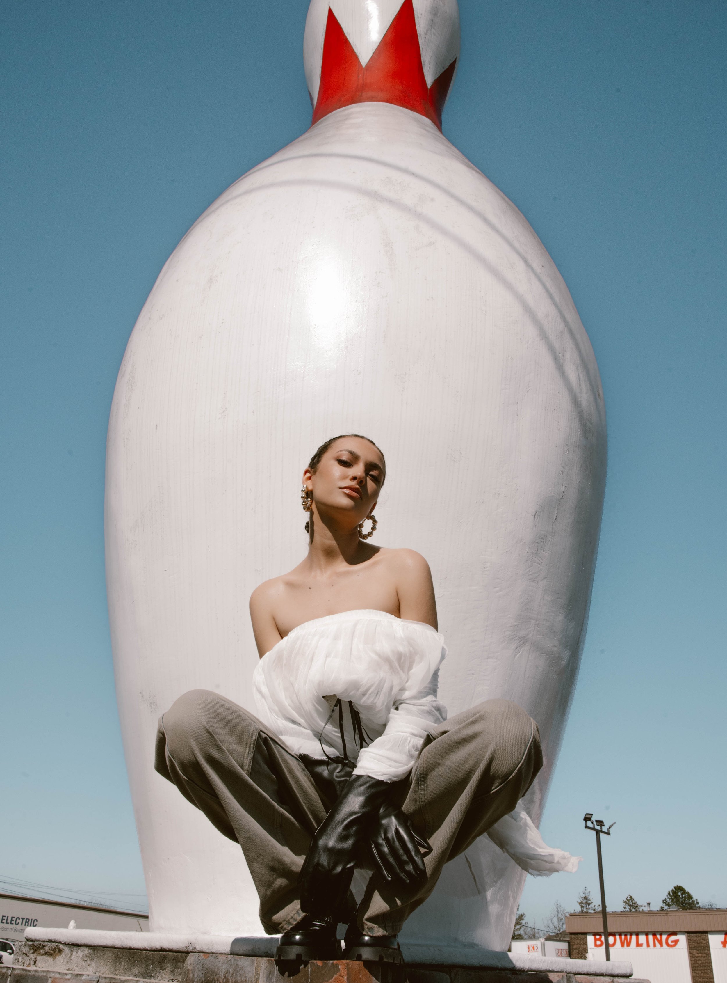 Woman poses near a large bowling pin sculpture wearing a white shawl and olive pants in an outdoor, sunny setting against a blue sky.