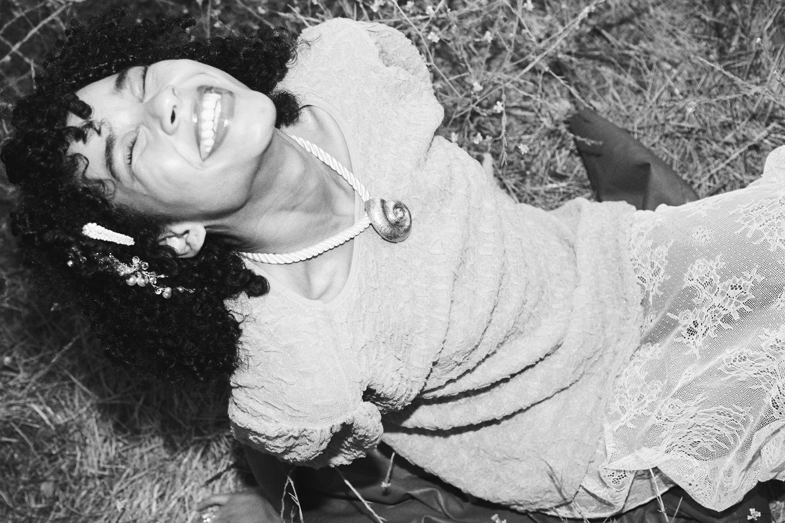 Monochrome portrait of an African American woman lying in a field, smiling with textured dress, statement necklace and hair accessory.