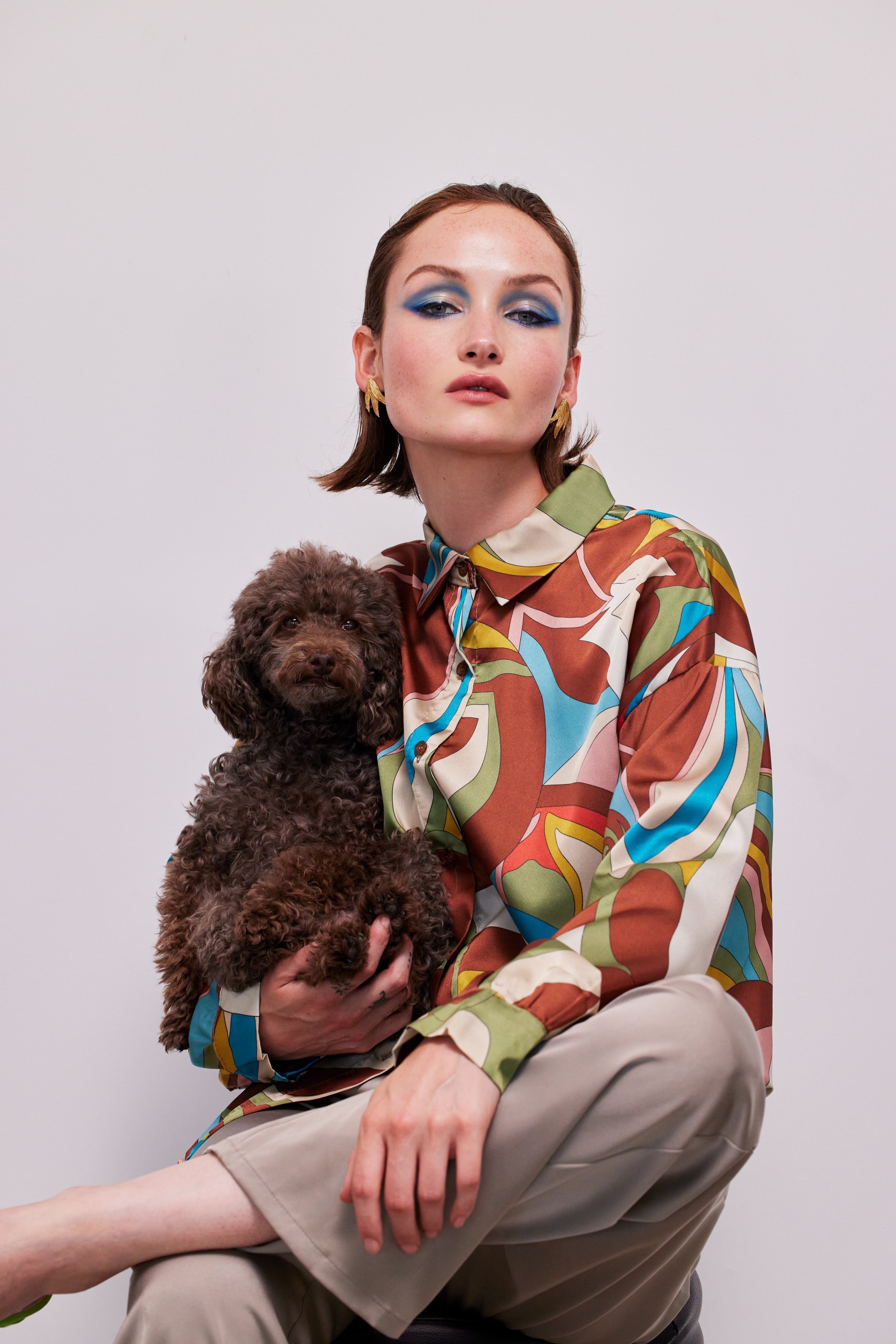 A woman with dramatic blue eye makeup and a patterned shirt holds a brown poodle, posing against a light background in an indoor setting.