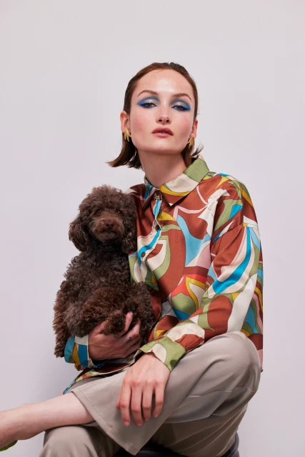 A woman with dramatic blue eye makeup and a patterned shirt holds a brown poodle, posing against a light background in an indoor setting.