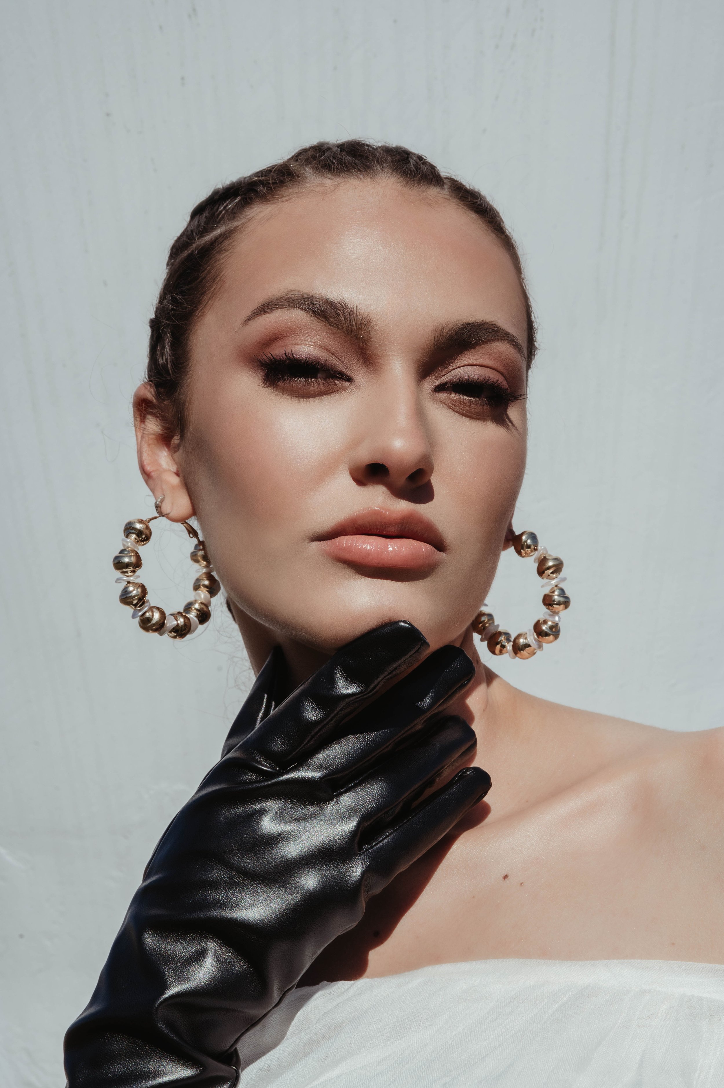 Close-up portrait of a young woman wearing gold hoop earrings and a black glove against a white background in a beauty shot.