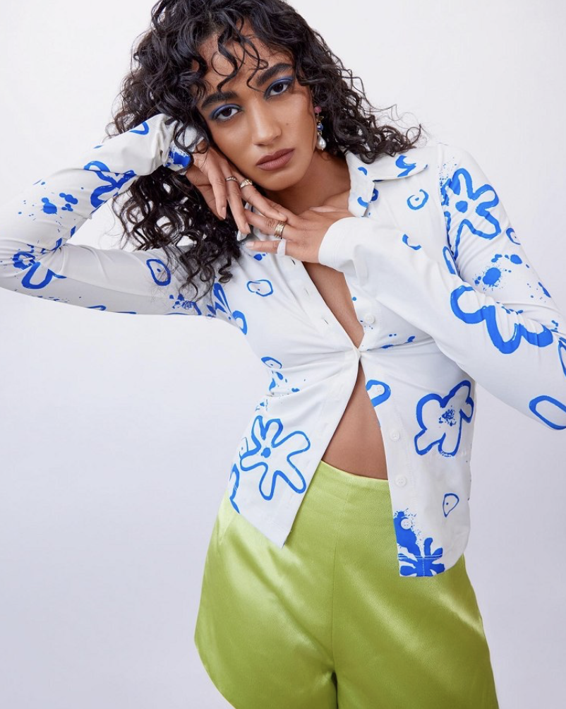 Fashionable young woman in a white button-down shirt with a blue pattern and green shorts posing in a studio with a neutral background.
