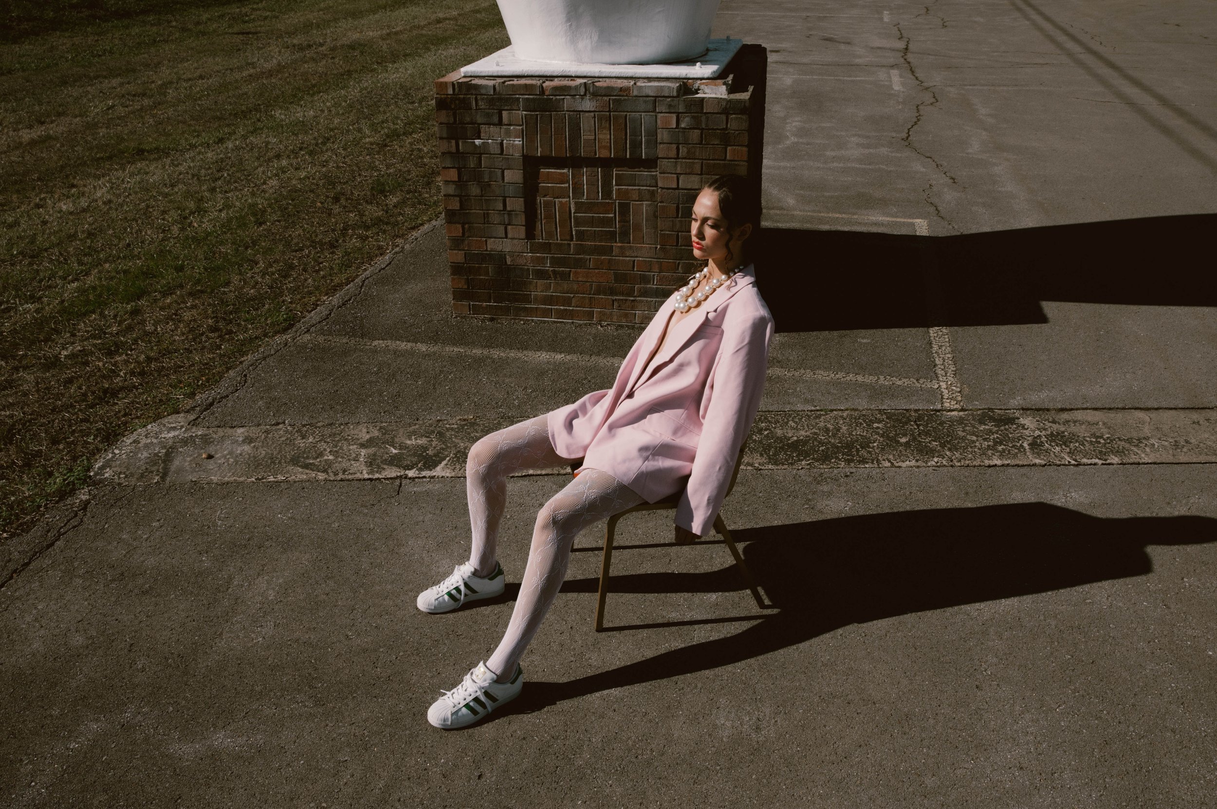 A stylish woman sits on a chair outdoors, wearing a pink blazer, white tights, sneakers, and a pearl necklace, basking in warm light.