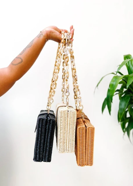 Three woven purses with chain straps in black, white, and tan are held by a hand against a plain white background with a plant.