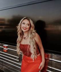 Blonde woman wearing a sparkling red sequin top and shiny red trousers leaning against a dark metallic vehicle.