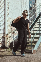 A young man wearing a patterned bucket hat, black sweater vest, and dark trousers standing by an outdoor metal staircase.