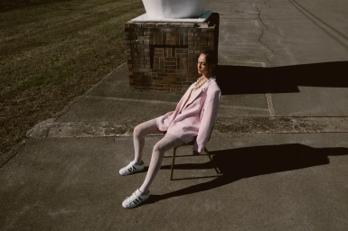Model seated on a chair outdoors wearing a pink blazer, white lace tights, and sneakers, styled by Kinsey Ranee.