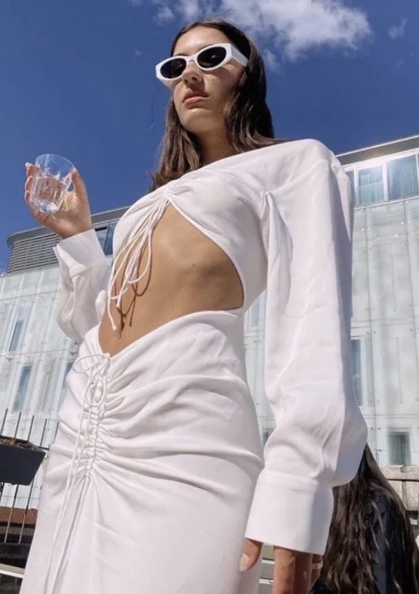 Woman in white cut-out dress and sunglasses holds a glass of water against a blue sky and modern building backdrop on a sunny day.