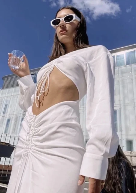 Woman in white cut-out dress and sunglasses holds a glass of water against a blue sky and modern building backdrop on a sunny day.