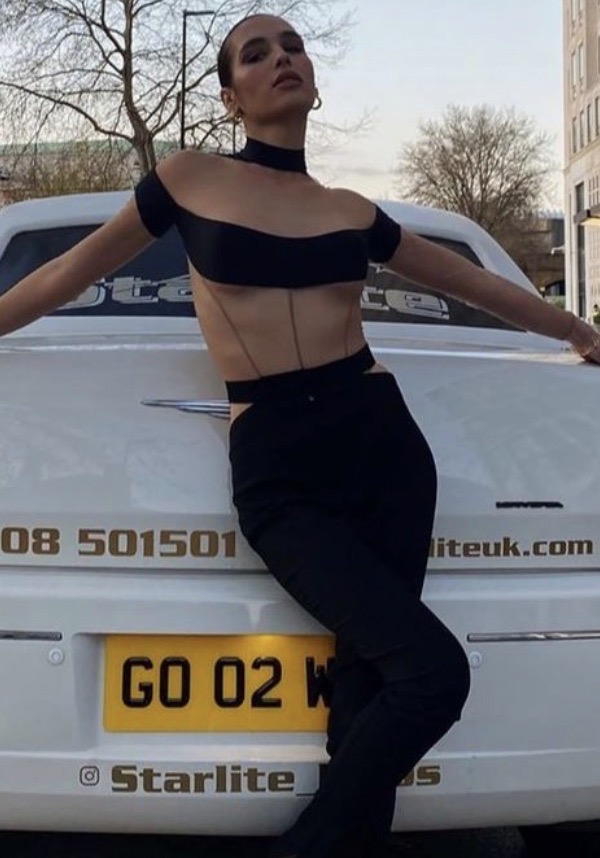 A young adult woman leans against a white car in black clothing, posing outdoors in an urban setting during the daytime.