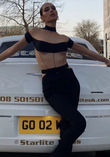 A young adult woman leans against a white car in black clothing, posing outdoors in an urban setting during the daytime.