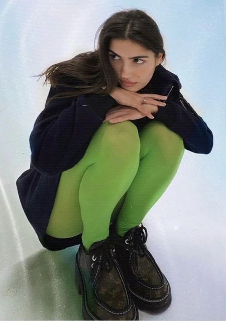 Brunette woman in a crouched pose wearing a dark jacket, bright green tights, and platform shoes sitting on a white reflective surface.
