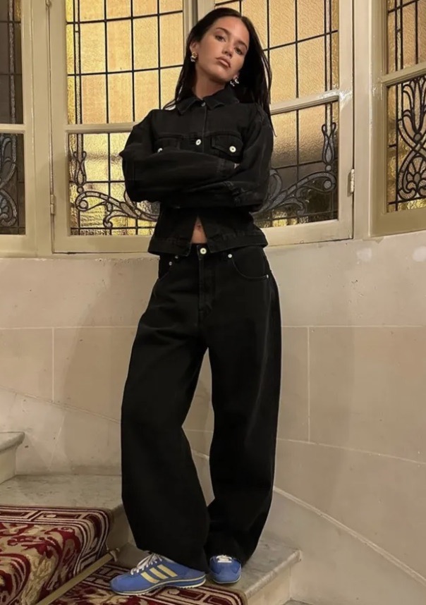 A woman in a black denim jacket and baggy jeans poses on stairs near a window with a grid pattern. The image emphasizes a fashion-forward look.