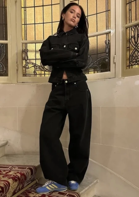 A woman in a black denim jacket and baggy jeans poses on stairs near a window with a grid pattern. The image emphasizes a fashion-forward look.