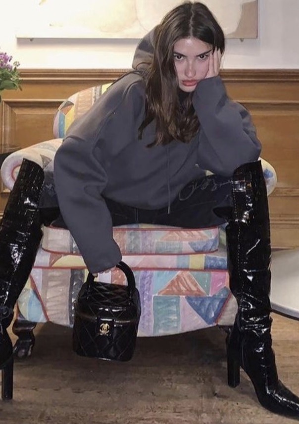 A young woman in a grey hoodie and black patent-leather boots poses in a colorful chair with a small, quilted Chanel handbag.