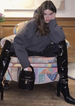 A young woman in a grey hoodie and black patent-leather boots poses in a colorful chair with a small, quilted Chanel handbag.