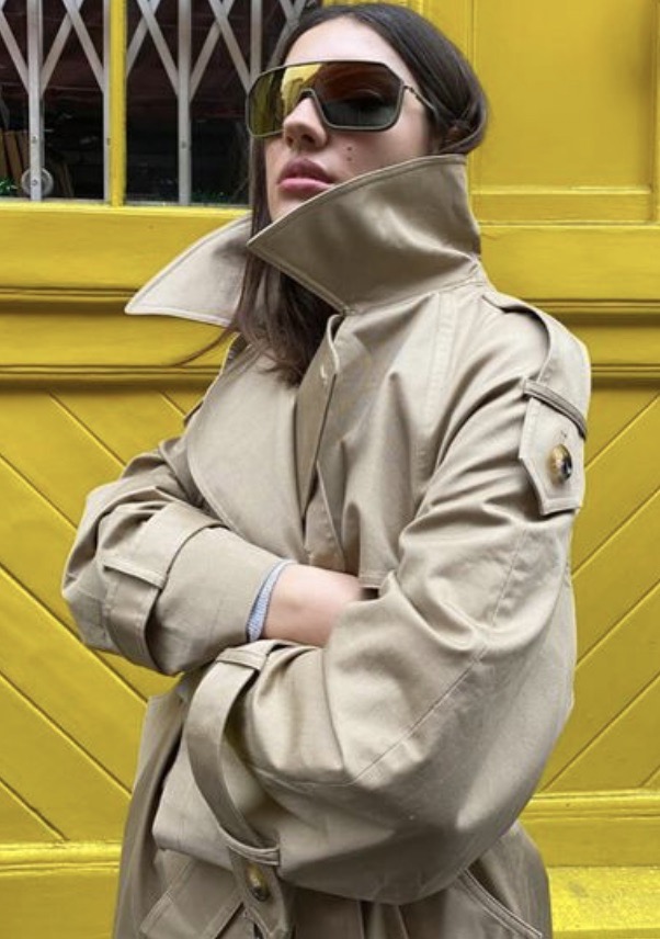 Woman poses in a trench coat and sunglasses against a yellow door. The medium shot captures the coat's details and the door's texture.