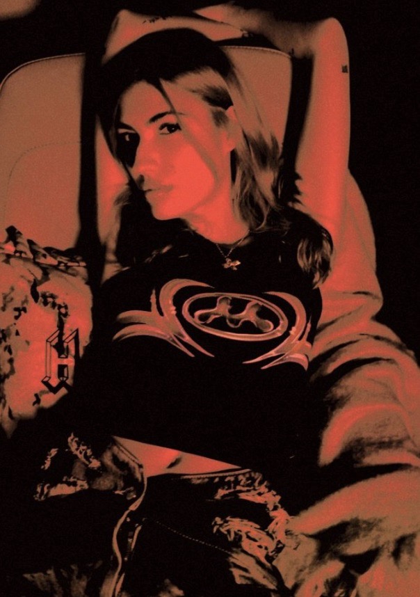 Woman in a black shirt with a graphic design reclining on a patterned chair in a low-light setting with a red color overlay.