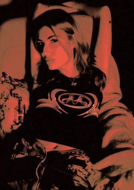 Woman in a black shirt with a graphic design reclining on a patterned chair in a low-light setting with a red color overlay.