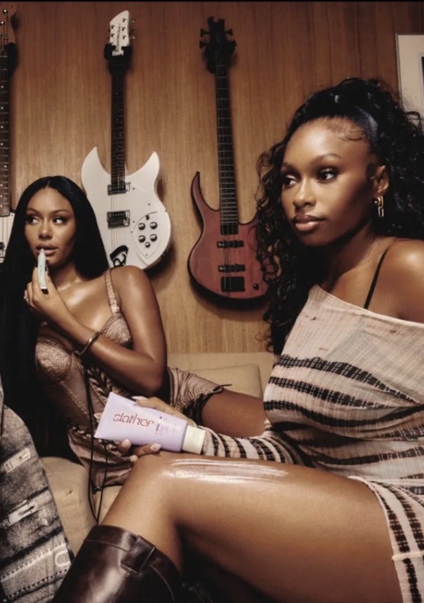 Two African-American women indoors in chic dresses and boots, one applying moisturizer, with guitars in the background, warm lighting.