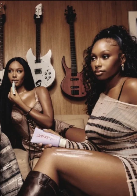Two African-American women indoors in chic dresses and boots, one applying moisturizer, with guitars in the background, warm lighting.